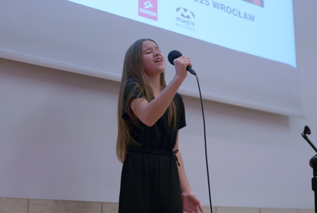 Dziewczynka w wieku około 10 lat śpiewająca do mikrofonu na tle banneru TAURON Music Star