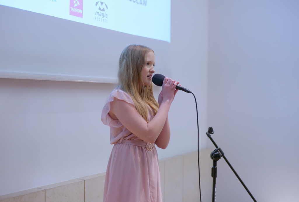 Dziewczynka w wieku około 10 lat śpiewająca do mikrofonu na tle banneru TAURON Music Star