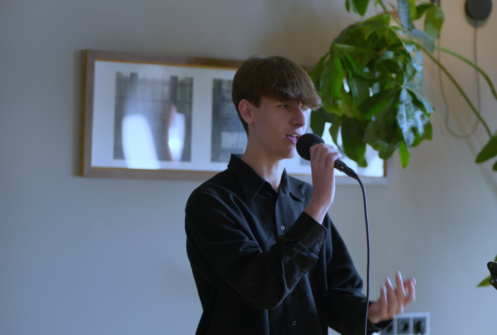 Chłopiec w wieku około 13 lat w czarnej koszuli śpiewający do mikrofonu na tle rośliny i obrazu 