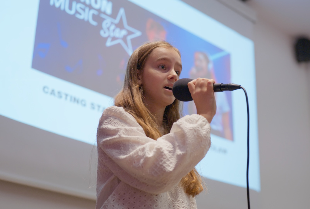 Dziewczynka w wieku około 9 lat śpiewająca do mikrofonu na tle banneru TAURON Music Star