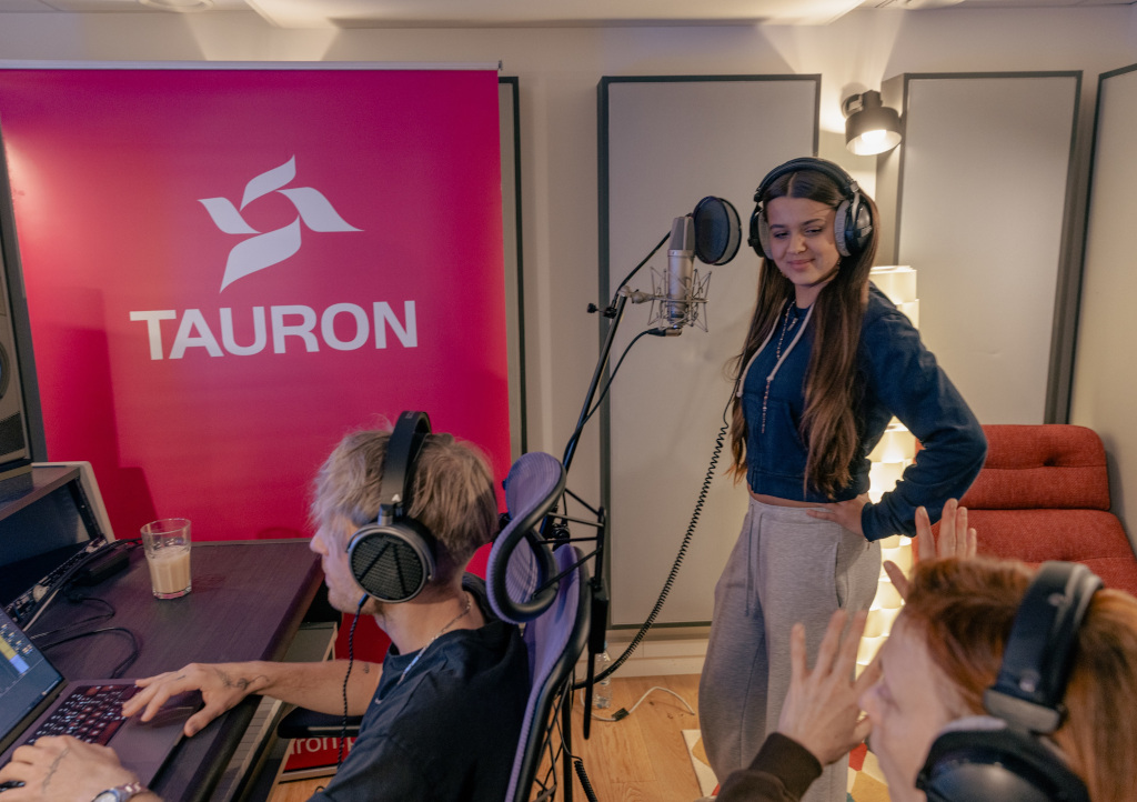 Studio nagraniowe, na pierwszym planie producent muzyczny, za nim uśmiechnięta dziewczyna przed mikrofonem